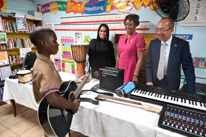 Deputy Prime Minister and Minister of National Security, the Hon. Dr. Horace Chang (right), Permanent Secretary, Ambassador Alison Stone Roofe (centre) and Vice Principal for Kingston High School, Sashauna Gordon (left) looks on as grade seven student, Kimani Davis strums one of the guitars handed over to the institution by the Ministry of National Security today (April 9).